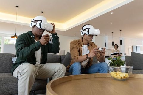 Friends playing VR games on livingroom sofa wearing white headsets holding controllers