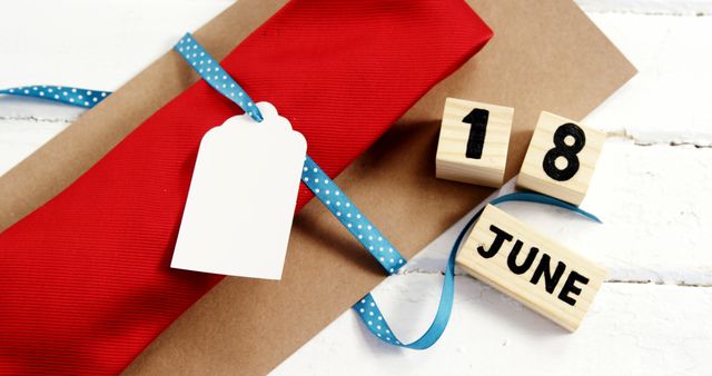 A red gift with a blank tag and a blue ribbon lies next to wooden blocks displaying the date 18 June, with copy space. This setup suggests preparation for a celebration or a reminder of an important date, Father's Day or a birthday.