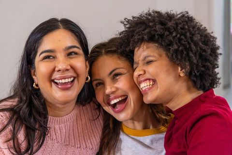 Three diverse women laughing and hugging, wearing knit sweaters and gold hoop earrings