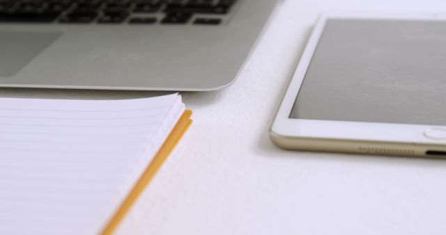 Close-up of a workplace setup including a laptop, tablet, and a notebook on a white desk. Useful for illustrating modern office environments, remote working situations, or educational contexts. Perfect for blogs, articles, and advertisements focused on productivity, technology, or workplace organization.