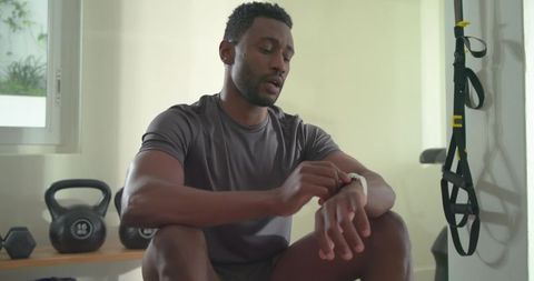 African american man checking smartwatch in home gym with kettlebell and suspension trainer