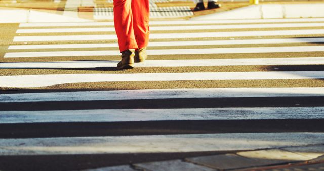 Person crossing street at marked pedestrian crosswalk wearing orange pants and black shoes. Ideal for illustrating themes of urban life, road safety, pedestrian crossing etiquette, city commuting, and transportation.