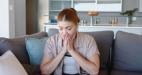 Woman covering mouth while leaning on gray sofa, expressing worry and quiet contemplation