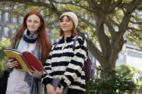 Multicultural female students standing on campus under tree, holding notebooks and tablet