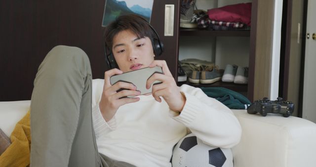 Asian boy wearing headphones playing game on smartphone sitting on the couch at home. teenager lifestyle and living concept