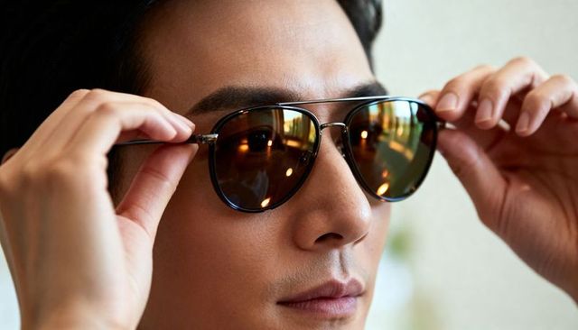 Asian man adjusting aviator sunglasses with amber reflective lenses and warm moody lighting