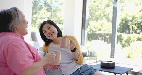 Two Women Laughing and Drinking Coffee on Sofa by Glass Doors, Sunlit Modern Living Space