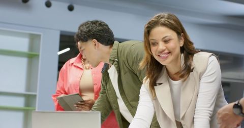 Diverse team leaning over laptop and tablet in modern office meeting, smiling woman leading