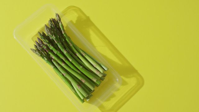 A collection of fresh asparagus stems neatly arranged in a clear plastic container, showcased against a lively yellow background. Perfect for use in food-related content such as grocery store advertisements, organic lifestyle blogs, or healthy eating campaigns. Highlights the freshness and nutritional benefits of asparagus as a takeaway dish or seasonal vegetable for balanced diets.