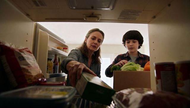 Mother reaching into fridge while son unpacking groceries from cardboard box in kitchen