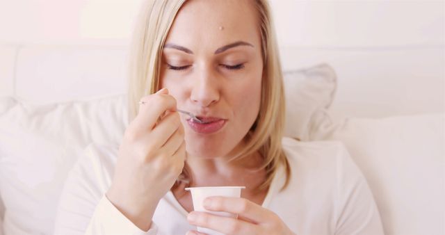 This image features a blonde woman sitting comfortably indoors and enjoying a container of yogurt. Ideal for use in advertisements for dairy products, health and wellness blogs, healthy lifestyle editorials, kitchen inspiration, and food-related content. Perfect for emphasizing themes of healthy eating, relaxation, and enjoyable meals.