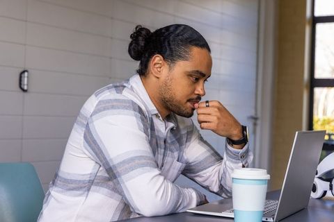Young man working on laptop in casual workspace with travel cup, smartwatch, headphones