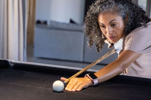 Senior african american woman aiming pool cue wearing smartwatch in modern home lounge