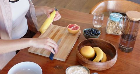 Hands slicing banana on wooden cutting board for healthy rustic breakfast prep with grapefruit