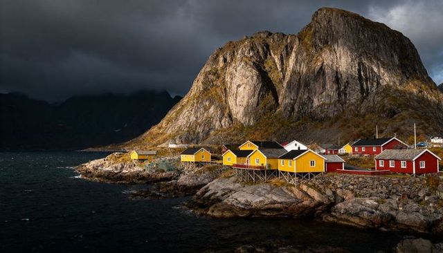 Sunlight striking yellow cabins on rocky fjord beneath dramatic granite cliff