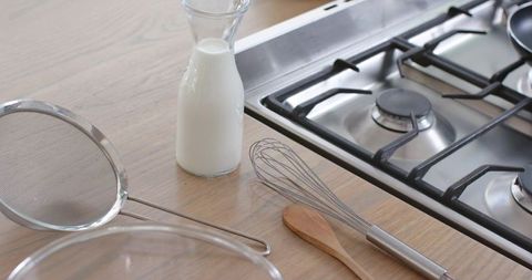 Glass milk bottle on light wood counter with whisk, strainer and gas cooktop for baking