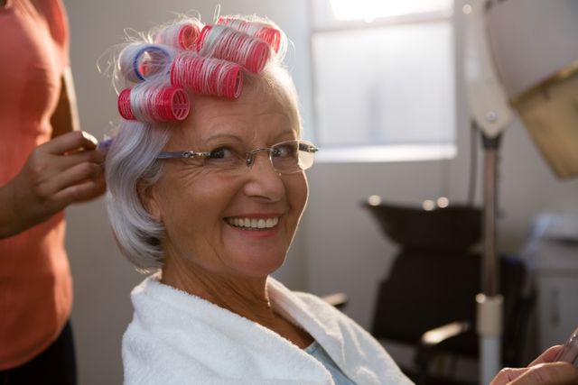 Senior woman sitting in a salon chair, smiling while a hairstylist removes curlers from her hair. Ideal for use in advertisements for beauty salons, hair care products, and services targeting elderly clients. Can also be used in articles about senior beauty tips and self-care.