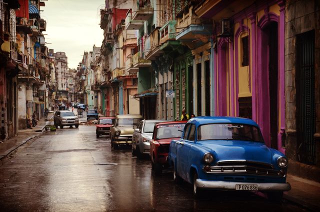 This image captures a nostalgic urban scene with vintage cars lined along a weathered street in a vibrant and colorful historical city, likely in Havana, Cuba. Ideal for travel agencies, blogs discussing vintage or cultural tours, history publications, or content focused on architecture and tourism. Ideal for conveying themes of nostalgia, cultural richness, and travel inspiration.