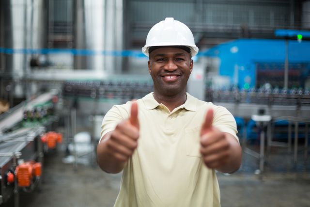 This image shows a cheerful factory worker giving a thumbs up in a beverage production plant. He is wearing a safety helmet and standing in front of industrial machinery. This photo can be used for promoting workplace safety, job satisfaction, industrial operations, and manufacturing processes. It is ideal for use in corporate materials, training manuals, and industry-related publications.