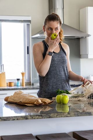 Young woman eating green apple in modern kitchen wearing smartwatch after workout