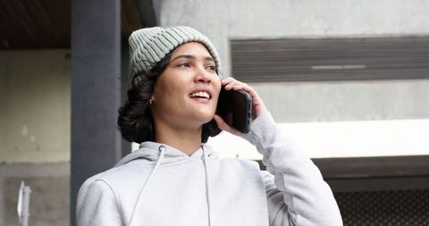 Young woman talking on smartphone under urban overhang wearing knit beanie and gray hoodie