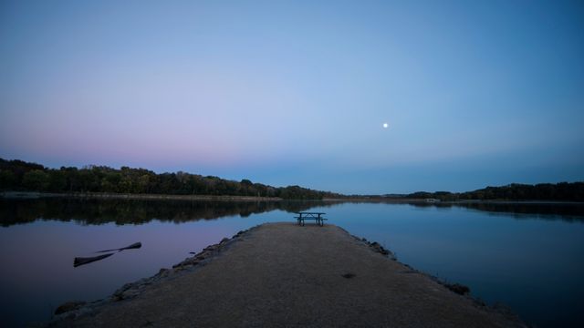 Photograph depicts a tranquil lake at dusk with the moon reflecting on the water. Nature setting shows a calm atmosphere with a clear sky and a serene landscape. This can be used for promoting relaxation, nature retreats, or outdoor activities.