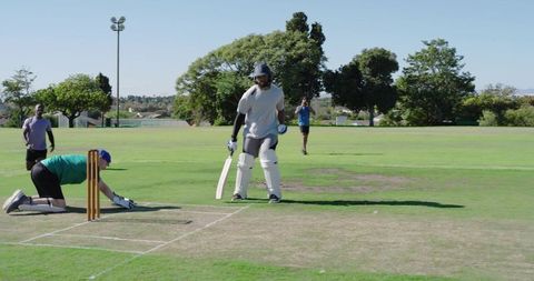 Batsman hitting cricket ball while wicketkeeper dives for run-out on sunny grass field