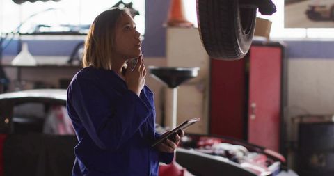 Female mechanic inspecting suspended wheel while holding tablet in busy auto repair garage bay