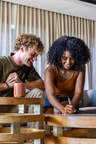Mixed-race couple leaning over coffee table writing paperwork while relaxing on sofa