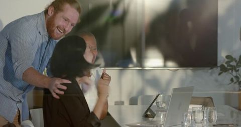 Laughing diverse team celebrating success around laptop in bright modern boardroom