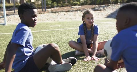 Diverse youth soccer teammates sitting on turf holding balls during sunny practice
