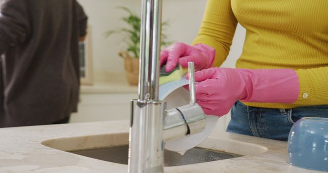 Woman in yellow top and jeans washing dishes in kitchen sink wearing pink gloves. Ideal for illustrating household chores, cleaning products, domestic tasks, and cleanliness themes.