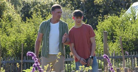 Father walking and talking with teenage son through sunlit allotment garden with flowers