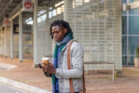 Mid adult african american commuter waiting at stop holding coffee and checking phone