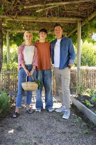 Family harvesting under pergola standing smiling with wicker basket in sunlit home garden