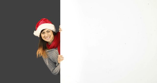 This image shows a woman with a smiling face happily peeking from behind a blank white space. She is dressed in a Santa hat and scarf, capturing the festive holiday spirit. This image can be used for Christmas and holiday-themed advertisements, greeting designs, or social media posts to promote seasonal joy and celebrations.