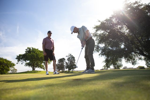 Two Caucasian male golfers practicing on a golf course on a sunny day wearing caps and golf clothes, holding golf clubs one man hitting a golf ball. Hobby healthy lifestyle leisure.