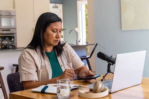 Mature asian woman recording podcast at home using laptop and smartphone