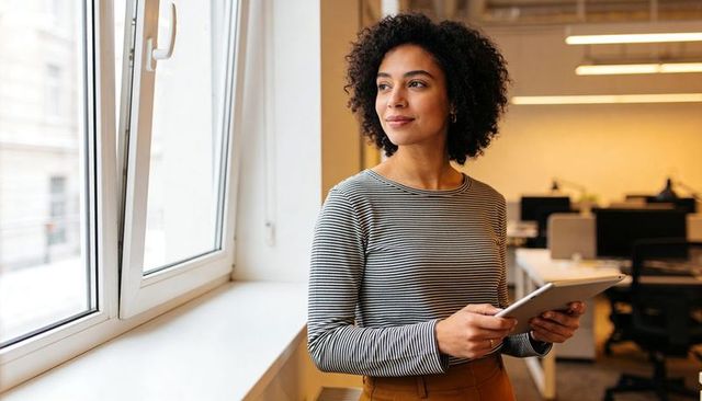 Professional woman holding tablet and gazing out window in modern open office workspace
