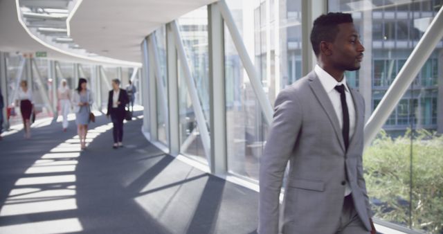 Business professional in grey suit walking confidently through a modern office corridor with large glass windows. Ideal for use in corporate presentations, business websites, and leadership promotions. Highlights the professional and contemporary work environment.