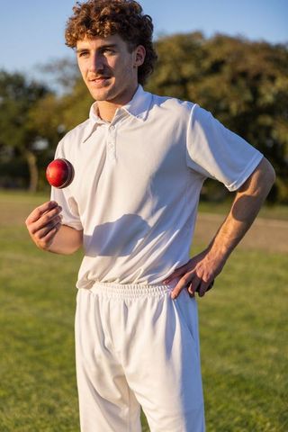 Young cricketer balancing red ball on finger wearing white kit on sunlit grassy pitch