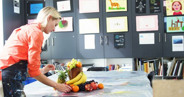 Art teacher in classroom arranging fruits for a still life setup. There are various colorful artworks displayed on the walls in the background. Ideal for use in educational materials, teaching resources, art school promotions, and creative learning concepts.