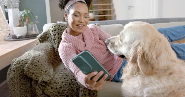 Woman reclining on sofa with a golden retriever, both looking at the phone screen. Warmly lit living room creates cozy atmosphere. Great for concepts of pet bonding, relaxed lifestyle, technology in casual settings, and home leisure activities.