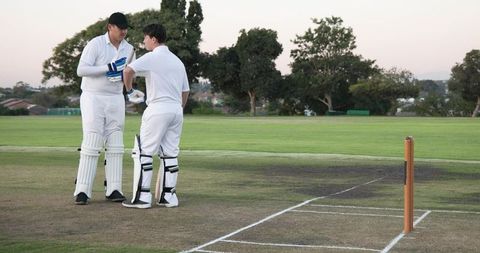Cricket teammates exchanging handshake on suburban pitch at dusk wearing white kit