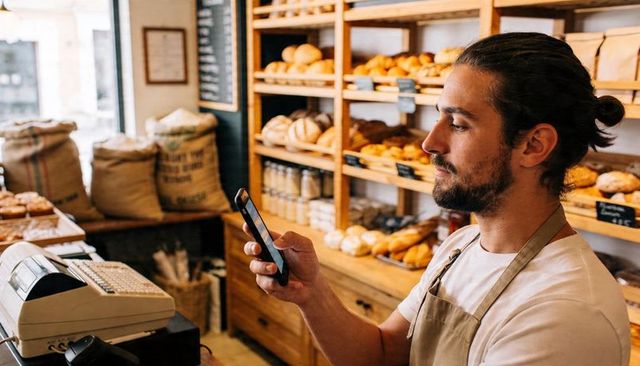 Baker checking smartphone orders inside rustic artisan bakery with shelves of fresh bread