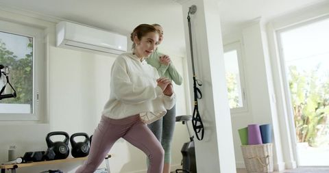 Two women lunging together in sunlit home gym wearing hooded sweatshirt and mauve leggings