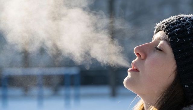 Woman exhaling visible breath vapor in snowy park wearing frosted knit beanie closeup