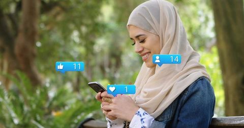 Young woman wearing hijab smiling while checking phone and getting social media likes