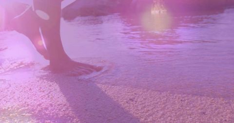 Barefoot man stepping into shallow pink-lit seawater on pebbly shore with lens flare