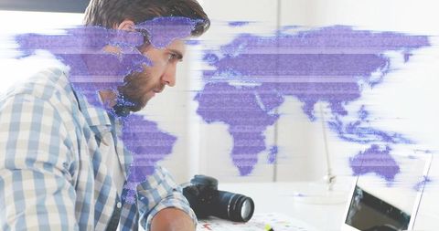 Bearded man leaning over laptop working with camera on desk and digital world map overlay